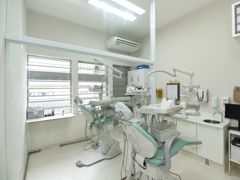 Sala de odontologia com as paredes brancas, uma janela ao fundo e duas cadeiras de dentista separadas por uma parede de vidro. De frente para as cadeiras fica a bancada com pia e instrumentos